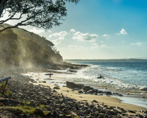 Sun shining through spray mist in the eucalyptus trees. Tropical stone pebbles in the foreground. Two surfers returning back to shore.